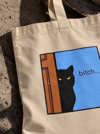Μπεζ tote bag με μαύρη γάτα με κίτρινα μάτια πίσω από πόρτα και τη φράση bitch