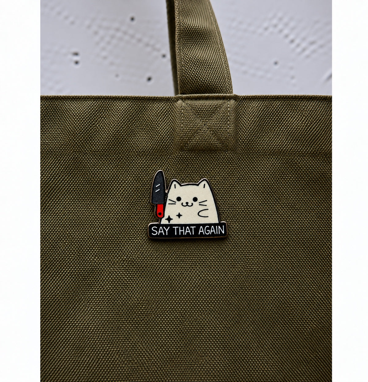 White cat with knife wooden printed pin saying Say That Again displayed on olive green bag