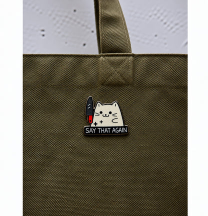 White cat with knife wooden printed pin saying Say That Again displayed on olive green bag